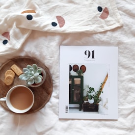 A white magazine cover with the title '91 Magazine' on a soft white blanket, alongside a wooden tray containing a cup of coffee, cookies, and a small succulent plant.