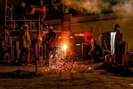Several workers are wearing protective gear in an industrial setting, engaging in metalworking or a foundry process. Molten metal pours from a furnace, emitting bright sparks and creating a warm glow. Equipment and structural elements surround them, and a haze lingers in the air.