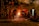 Several workers are wearing protective gear in an industrial setting, engaging in metalworking or a foundry process. Molten metal pours from a furnace, emitting bright sparks and creating a warm glow. Equipment and structural elements surround them, and a haze lingers in the air.