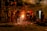 Several workers are wearing protective gear in an industrial setting, engaging in metalworking or a foundry process. Molten metal pours from a furnace, emitting bright sparks and creating a warm glow. Equipment and structural elements surround them, and a haze lingers in the air.