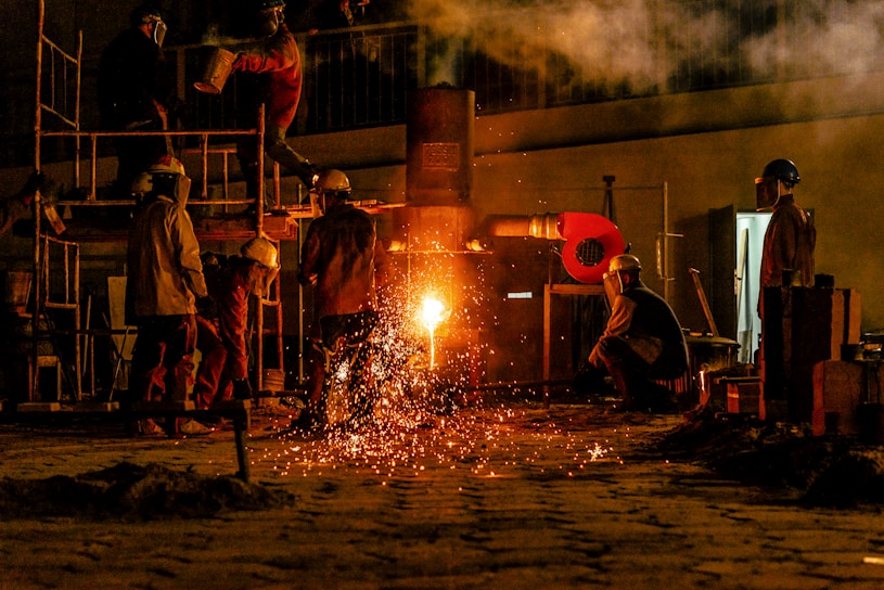 Several workers are wearing protective gear in an industrial setting, engaging in metalworking or a foundry process. Molten metal pours from a furnace, emitting bright sparks and creating a warm glow. Equipment and structural elements surround them, and a haze lingers in the air.