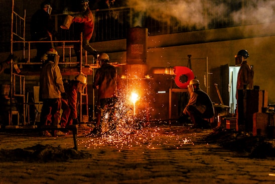 Several workers are wearing protective gear in an industrial setting, engaging in metalworking or a foundry process. Molten metal pours from a furnace, emitting bright sparks and creating a warm glow. Equipment and structural elements surround them, and a haze lingers in the air.