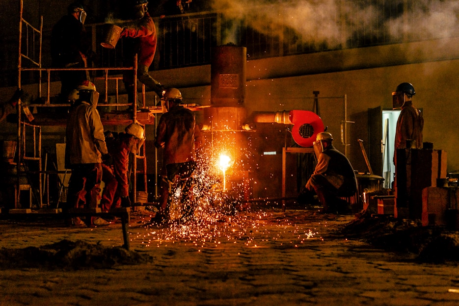 Several workers are wearing protective gear in an industrial setting, engaging in metalworking or a foundry process. Molten metal pours from a furnace, emitting bright sparks and creating a warm glow. Equipment and structural elements surround them, and a haze lingers in the air.