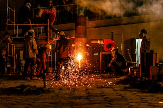 Several workers are wearing protective gear in an industrial setting, engaging in metalworking or a foundry process. Molten metal pours from a furnace, emitting bright sparks and creating a warm glow. Equipment and structural elements surround them, and a haze lingers in the air.
