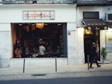A bar or café with an open-front design features several people seated inside, engaging in conversation and looking towards the street. The sign above the entrance reads 'Crafty Corner' in neon lights. A chandelier hanging from the ceiling adds a warm ambiance. A man walks past on the sidewalk, in front of the establishment, where another sign advertises a different brand.