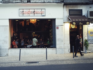 A bar or café with an open-front design features several people seated inside, engaging in conversation and looking towards the street. The sign above the entrance reads 'Crafty Corner' in neon lights. A chandelier hanging from the ceiling adds a warm ambiance. A man walks past on the sidewalk, in front of the establishment, where another sign advertises a different brand.