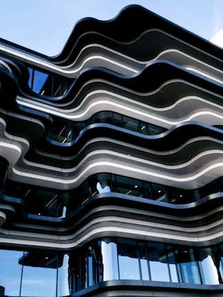 The image features a modern architectural building with a unique wavy design. The structure is composed of multiple layers with undulating lines, creating a dynamic and visually appealing façade. Large glass windows interspersed throughout the building reflect the sky, adding to the sleek and contemporary feel.