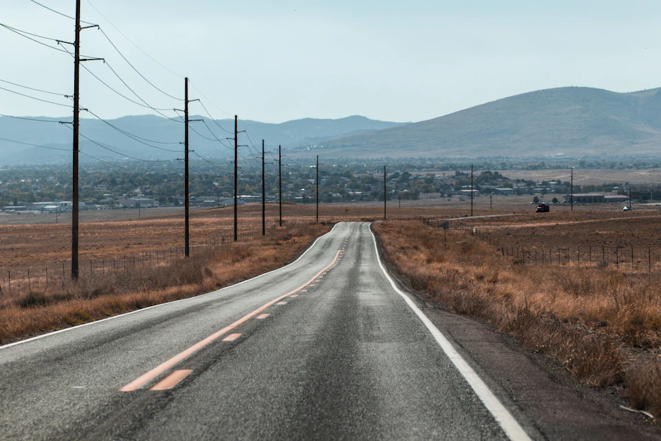 The image depicts a long, straight road cutting through a dry, desert-like landscape. The road is marked with white lines and stretches out into the distance, disappearing beyond the horizon. Power lines run parallel to the road on both sides, adding an industrial touch to the otherwise barren surroundings. In the background, distant mountains rise against the sky, their peaks shrouded in mist.