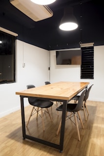 A modern conference room features a wooden table with a black metal frame, surrounded by four black chairs with wooden legs. The room has wooden flooring and white walls with a dark upper section. The ceiling has a large hanging light fixture, and there is a wall-mounted air conditioning unit and a flat-screen monitor.