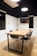 A modern conference room features a wooden table with a black metal frame, surrounded by four black chairs with wooden legs. The room has wooden flooring and white walls with a dark upper section. The ceiling has a large hanging light fixture, and there is a wall-mounted air conditioning unit and a flat-screen monitor.