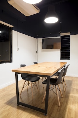A modern conference room features a wooden table with a black metal frame, surrounded by four black chairs with wooden legs. The room has wooden flooring and white walls with a dark upper section. The ceiling has a large hanging light fixture, and there is a wall-mounted air conditioning unit and a flat-screen monitor.