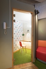 A small, enclosed office booth with a glass door containing colorful dots on the interior wall. Inside, there is a wooden table with a red chair on green carpet flooring. The booth is lit by a small overhead lamp, and there is a phone mounted on the adjacent exterior wall. The surrounding space includes a modern seating area with a section of a red and orange cushioned bench.