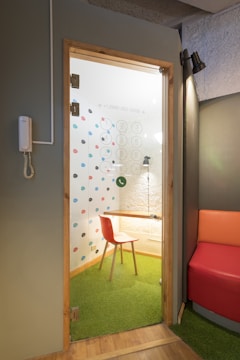 A small, enclosed office booth with a glass door containing colorful dots on the interior wall. Inside, there is a wooden table with a red chair on green carpet flooring. The booth is lit by a small overhead lamp, and there is a phone mounted on the adjacent exterior wall. The surrounding space includes a modern seating area with a section of a red and orange cushioned bench.