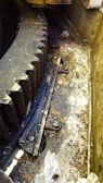 A large industrial gear with visible teeth, surrounded by a greasy and dirty environment, marked by scattered debris and grime. The area suggests heavy use and maintenance requirements, possibly within a mechanical or manufacturing setting. Metal components and bolts are also prominent.