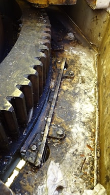 A large industrial gear with visible teeth, surrounded by a greasy and dirty environment, marked by scattered debris and grime. The area suggests heavy use and maintenance requirements, possibly within a mechanical or manufacturing setting. Metal components and bolts are also prominent.