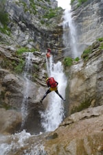 Two climbers are navigating a challenging waterfall descent in a rocky canyon. They are using ropes and wearing helmets and backpacks. The scene is surrounded by steep rock walls covered with greenery, and water cascades powerfully down the rocks.