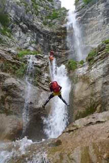 Two climbers are navigating a challenging waterfall descent in a rocky canyon. They are using ropes and wearing helmets and backpacks. The scene is surrounded by steep rock walls covered with greenery, and water cascades powerfully down the rocks.