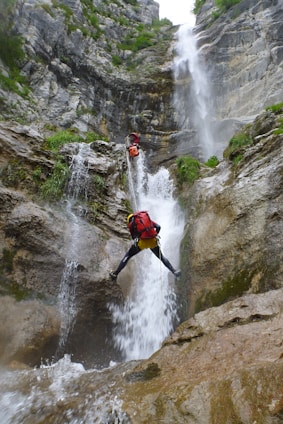 Two climbers are navigating a challenging waterfall descent in a rocky canyon. They are using ropes and wearing helmets and backpacks. The scene is surrounded by steep rock walls covered with greenery, and water cascades powerfully down the rocks.