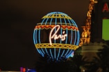 A brightly lit neon globe with the word 'Paris' in cursive, featuring blue, red, and yellow colors. It is surrounded by a partial view of the Eiffel Tower structure and some buildings in the background.