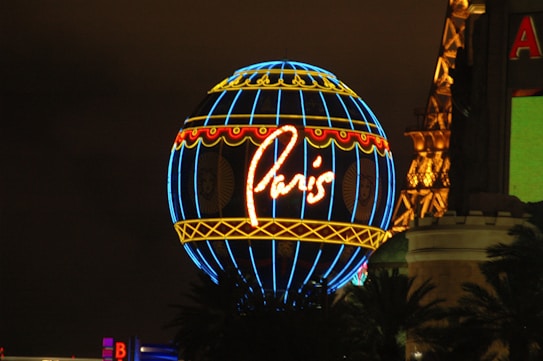 A brightly lit neon globe with the word 'Paris' in cursive, featuring blue, red, and yellow colors. It is surrounded by a partial view of the Eiffel Tower structure and some buildings in the background.