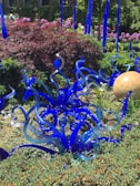 A vibrant garden features intricate blue glass sculptures that resemble plant tendrils amidst lush greenery and reddish-purple bushes. In the background, a spherical orange glass object sits among the foliage, with clusters of blooming pink flowers adding color to the scene.