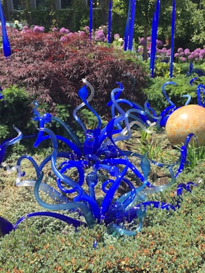 A vibrant garden features intricate blue glass sculptures that resemble plant tendrils amidst lush greenery and reddish-purple bushes. In the background, a spherical orange glass object sits among the foliage, with clusters of blooming pink flowers adding color to the scene.