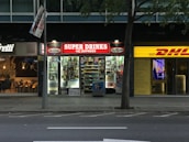 The image depicts a nighttime street view showing three adjacent stores. The central store has a bright red illuminated sign that reads 'SUPER DRINKS EXPRESS.' The store's interior is well-lit, showcasing shelves stocked with various drinks and snacks. To the left of this store is a café with a warmer, cozier ambiance, featuring hanging lights and visible seating arrangements. On the right side, there is a DHL express office with a bright yellow sign and an illuminated interior.