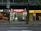 The image depicts a nighttime street view showing three adjacent stores. The central store has a bright red illuminated sign that reads 'SUPER DRINKS EXPRESS.' The store's interior is well-lit, showcasing shelves stocked with various drinks and snacks. To the left of this store is a café with a warmer, cozier ambiance, featuring hanging lights and visible seating arrangements. On the right side, there is a DHL express office with a bright yellow sign and an illuminated interior.