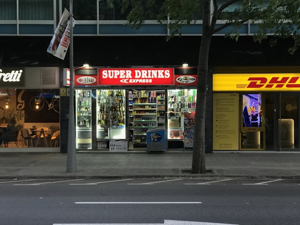 The image depicts a nighttime street view showing three adjacent stores. The central store has a bright red illuminated sign that reads 'SUPER DRINKS EXPRESS.' The store's interior is well-lit, showcasing shelves stocked with various drinks and snacks. To the left of this store is a café with a warmer, cozier ambiance, featuring hanging lights and visible seating arrangements. On the right side, there is a DHL express office with a bright yellow sign and an illuminated interior.
