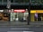 The image depicts a nighttime street view showing three adjacent stores. The central store has a bright red illuminated sign that reads 'SUPER DRINKS EXPRESS.' The store's interior is well-lit, showcasing shelves stocked with various drinks and snacks. To the left of this store is a café with a warmer, cozier ambiance, featuring hanging lights and visible seating arrangements. On the right side, there is a DHL express office with a bright yellow sign and an illuminated interior.