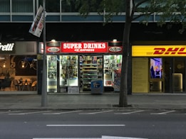 The image depicts a nighttime street view showing three adjacent stores. The central store has a bright red illuminated sign that reads 'SUPER DRINKS EXPRESS.' The store's interior is well-lit, showcasing shelves stocked with various drinks and snacks. To the left of this store is a café with a warmer, cozier ambiance, featuring hanging lights and visible seating arrangements. On the right side, there is a DHL express office with a bright yellow sign and an illuminated interior.