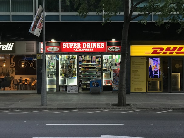 The image depicts a nighttime street view showing three adjacent stores. The central store has a bright red illuminated sign that reads 'SUPER DRINKS EXPRESS.' The store's interior is well-lit, showcasing shelves stocked with various drinks and snacks. To the left of this store is a caf&eacute; with a warmer, cozier ambiance, featuring hanging lights and visible seating arrangements. On the right side, there is a DHL express office with a bright yellow sign and an illuminated interior.