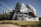A modern, innovative building characterized by its undulating, metallic facade which gives it a dynamic and fluid appearance. The structure features numerous windows and is set against a clear blue sky, surrounded by manicured grassy areas and trees.