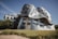 A modern, innovative building characterized by its undulating, metallic facade which gives it a dynamic and fluid appearance. The structure features numerous windows and is set against a clear blue sky, surrounded by manicured grassy areas and trees.