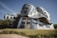 A modern, innovative building characterized by its undulating, metallic facade which gives it a dynamic and fluid appearance. The structure features numerous windows and is set against a clear blue sky, surrounded by manicured grassy areas and trees.