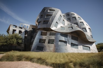 A modern, innovative building characterized by its undulating, metallic facade which gives it a dynamic and fluid appearance. The structure features numerous windows and is set against a clear blue sky, surrounded by manicured grassy areas and trees.