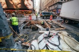 Several construction workers are engaged in roadwork in a busy urban area. Portable orange barriers and caution tape surround the work site. There are various construction materials, such as wooden planks, pipes, and large bags, scattered around. A couple of workers are inside a dug-up section of the ground. Surrounding the construction zone are high-rise buildings with commercial advertisements, vehicles including a truck and a SUV, and a bustling street environment suggesting a city setting.