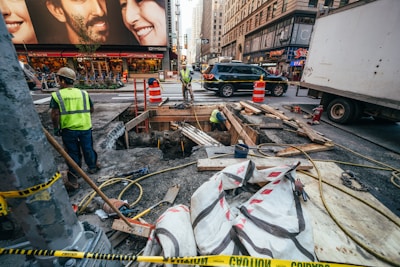 Several construction workers are engaged in roadwork in a busy urban area. Portable orange barriers and caution tape surround the work site. There are various construction materials, such as wooden planks, pipes, and large bags, scattered around. A couple of workers are inside a dug-up section of the ground. Surrounding the construction zone are high-rise buildings with commercial advertisements, vehicles including a truck and a SUV, and a bustling street environment suggesting a city setting.