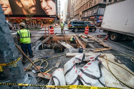 Several construction workers are engaged in roadwork in a busy urban area. Portable orange barriers and caution tape surround the work site. There are various construction materials, such as wooden planks, pipes, and large bags, scattered around. A couple of workers are inside a dug-up section of the ground. Surrounding the construction zone are high-rise buildings with commercial advertisements, vehicles including a truck and a SUV, and a bustling street environment suggesting a city setting.