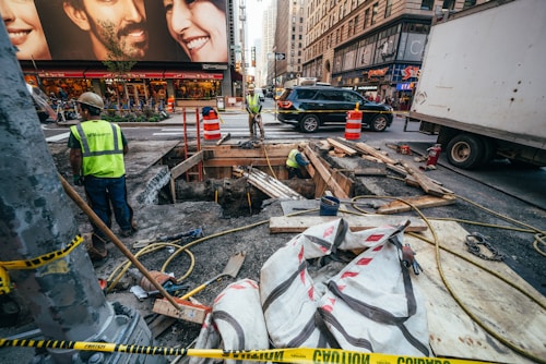 Several construction workers are engaged in roadwork in a busy urban area. Portable orange barriers and caution tape surround the work site. There are various construction materials, such as wooden planks, pipes, and large bags, scattered around. A couple of workers are inside a dug-up section of the ground. Surrounding the construction zone are high-rise buildings with commercial advertisements, vehicles including a truck and a SUV, and a bustling street environment suggesting a city setting.