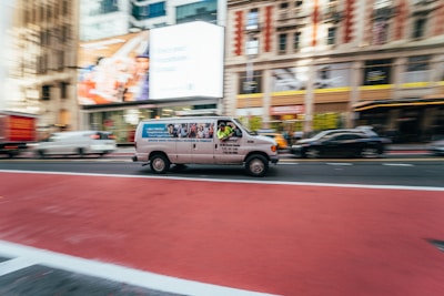 A white van with company advertisements on its side is moving quickly along a city street. The scene captures a sense of speed and motion, with a blurred background of office buildings and other vehicles. The road is marked with red and white lines, indicating a bus or special lane.