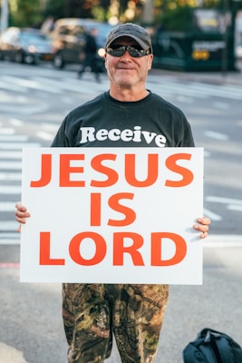 A man wearing sunglasses, a cap, and a black shirt with the word 'Receive' printed on it holds a large white sign with bold red text reading 'JESUS IS LORD' while standing on a street corner.