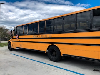 A large yellow school bus is parked on a concrete surface under a partly cloudy sky. The bus has a red stop sign extended from its side and is marked with black lettering indicating it belongs to a bus company.