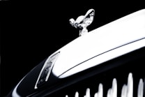 A close-up of a luxury car's hood ornament, featuring a sleek metallic emblem with intricate detailing. The background is dark, highlighting the shiny, reflective surface of the ornament.