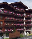 A wooden lodge with multiple balconies adorned with vibrant pink, purple, and white flowers. The building is made of dark wood with a rustic architectural style. There is a large wooden sign near the entrance indicating the name of the lodge. The setting appears to be serene and picturesque, likely in a mountainous region.