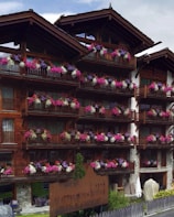 A wooden lodge with multiple balconies adorned with vibrant pink, purple, and white flowers. The building is made of dark wood with a rustic architectural style. There is a large wooden sign near the entrance indicating the name of the lodge. The setting appears to be serene and picturesque, likely in a mountainous region.