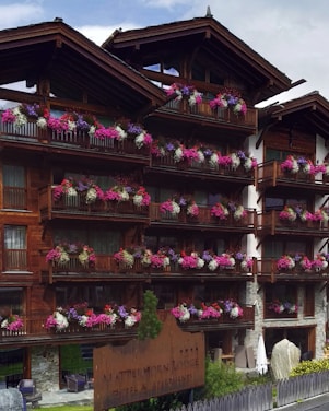A wooden lodge with multiple balconies adorned with vibrant pink, purple, and white flowers. The building is made of dark wood with a rustic architectural style. There is a large wooden sign near the entrance indicating the name of the lodge. The setting appears to be serene and picturesque, likely in a mountainous region.