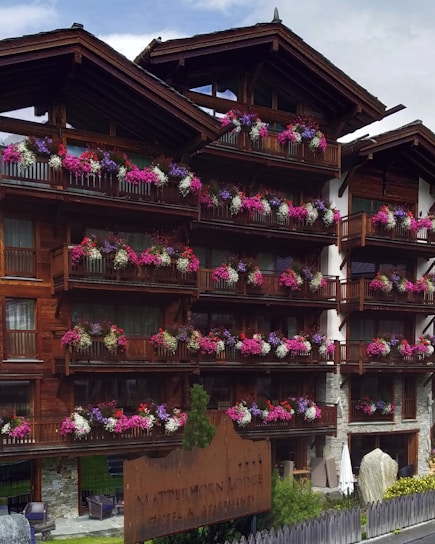 A wooden lodge with multiple balconies adorned with vibrant pink, purple, and white flowers. The building is made of dark wood with a rustic architectural style. There is a large wooden sign near the entrance indicating the name of the lodge. The setting appears to be serene and picturesque, likely in a mountainous region.