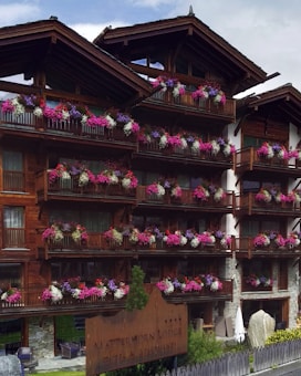 A wooden lodge with multiple balconies adorned with vibrant pink, purple, and white flowers. The building is made of dark wood with a rustic architectural style. There is a large wooden sign near the entrance indicating the name of the lodge. The setting appears to be serene and picturesque, likely in a mountainous region.