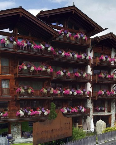 A wooden lodge with multiple balconies adorned with vibrant pink, purple, and white flowers. The building is made of dark wood with a rustic architectural style. There is a large wooden sign near the entrance indicating the name of the lodge. The setting appears to be serene and picturesque, likely in a mountainous region.